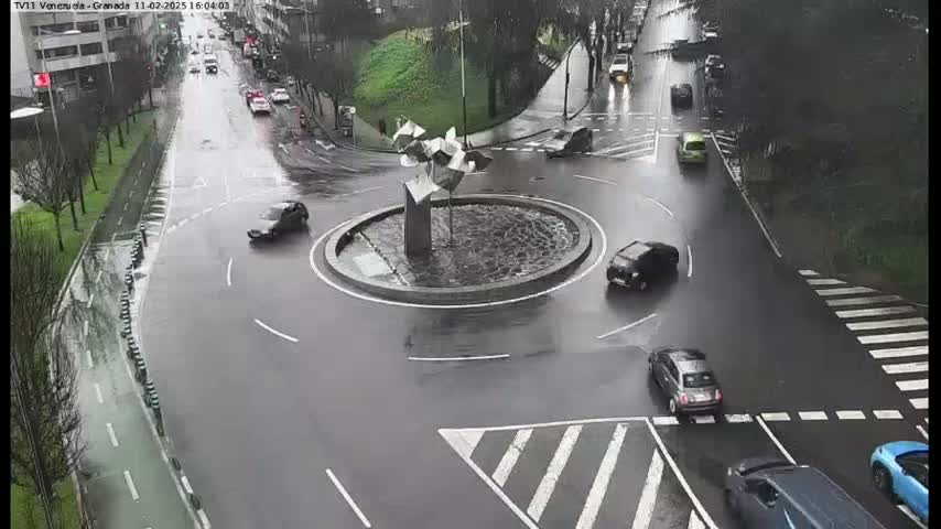 A wet roundabout with several cars navigating it, surrounded by roadways and greenery under a rainy sky.