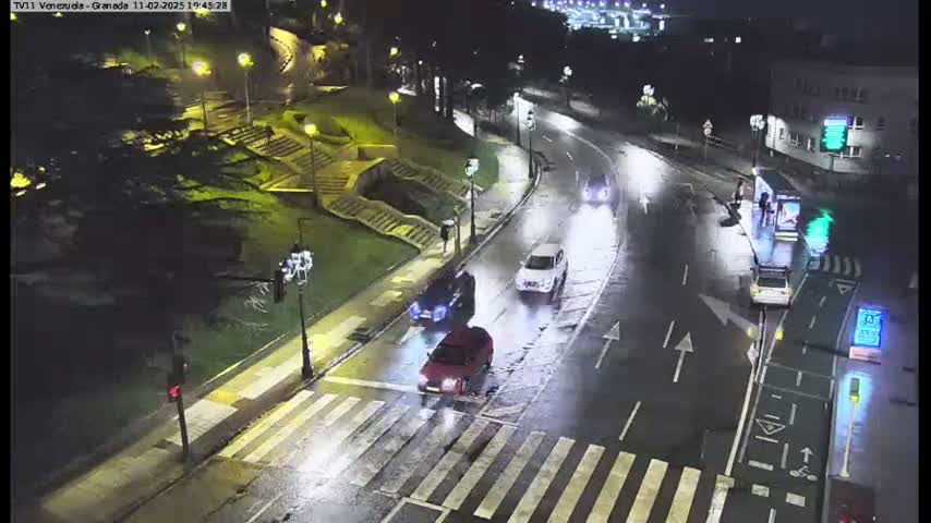 A wet, nighttime street scene shows several cars driving on a multi-lane road that intersects with a crosswalk, near some stairs and a bus stop.