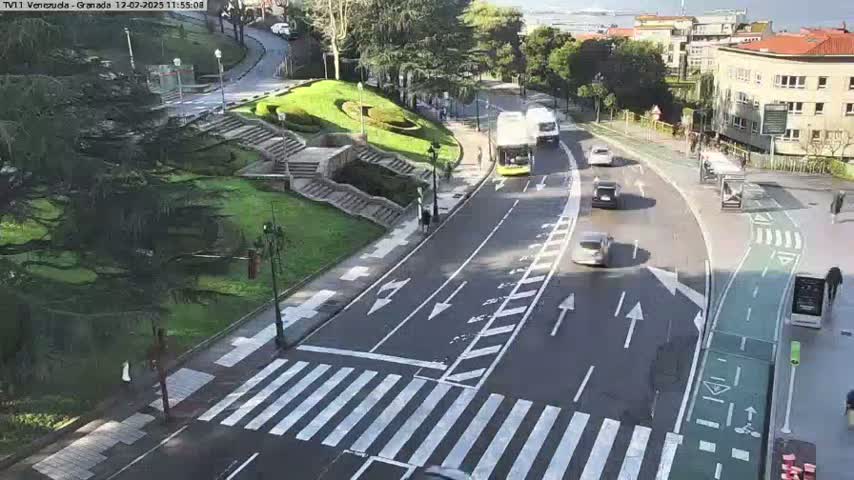 A sunny day reveals a roadway with several vehicles, including buses, cars, and a pedestrian crossing, alongside a landscaped area with stairs and greenery.