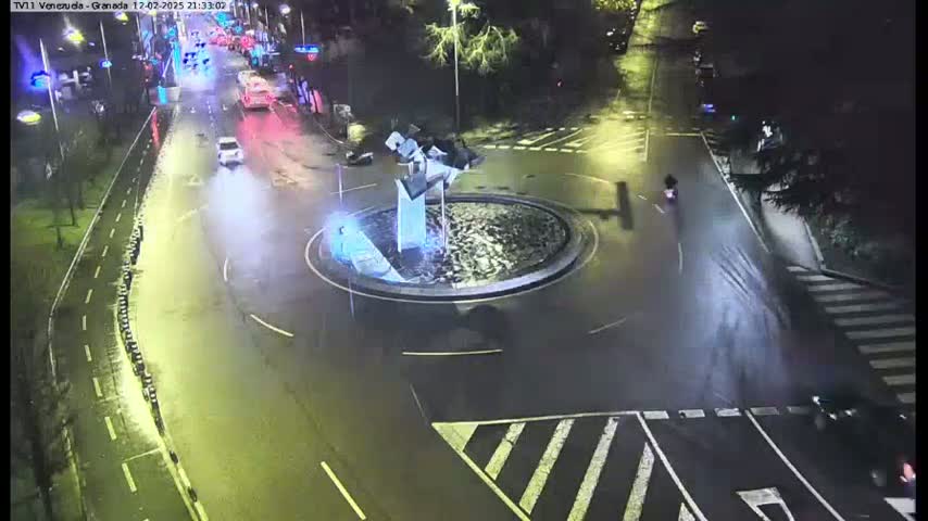 A wet, nighttime street scene shows vehicles traveling around a traffic circle that contains a modern sculpture.
