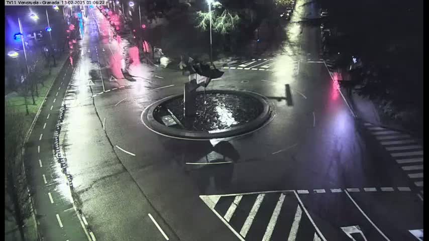 A wet roundabout at night is centered in the image, with roads and crosswalks leading away from it, and some vehicle lights visible in the distance.