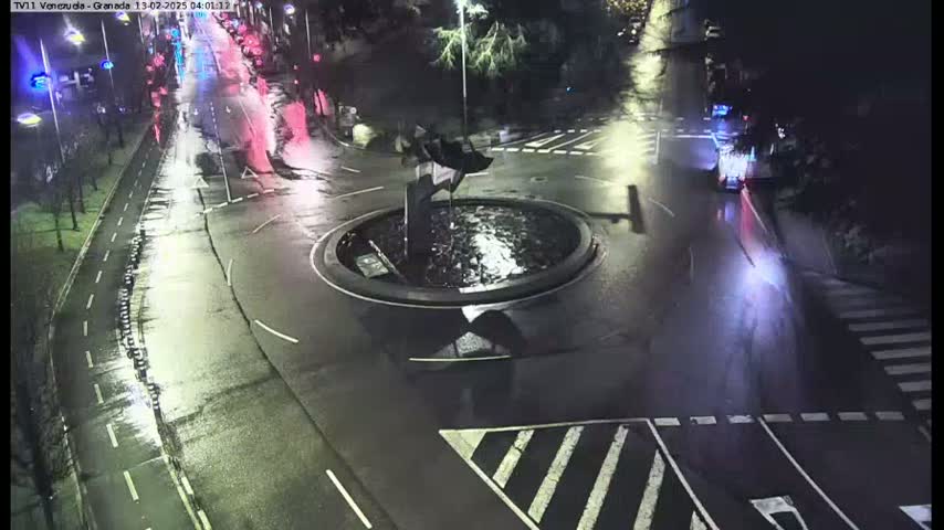 A wet, nighttime scene shows a roundabout with a central fountain and sparse traffic on surrounding roads.