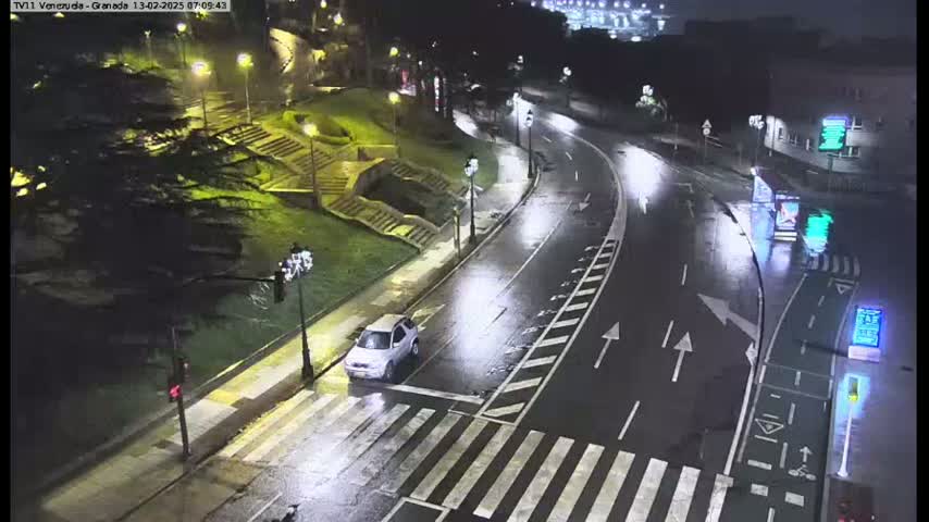A wet, curving road at night is seen from above, showing a pedestrian crossing with one car stopped at it and streetlights illuminating the scene.