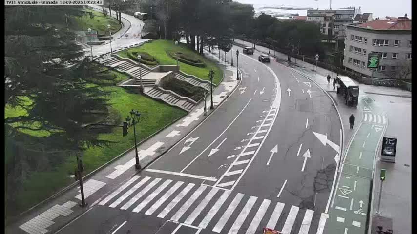 A wet road curves through a city park, past stairs and a bus stop, with a few pedestrians and vehicles visible.
