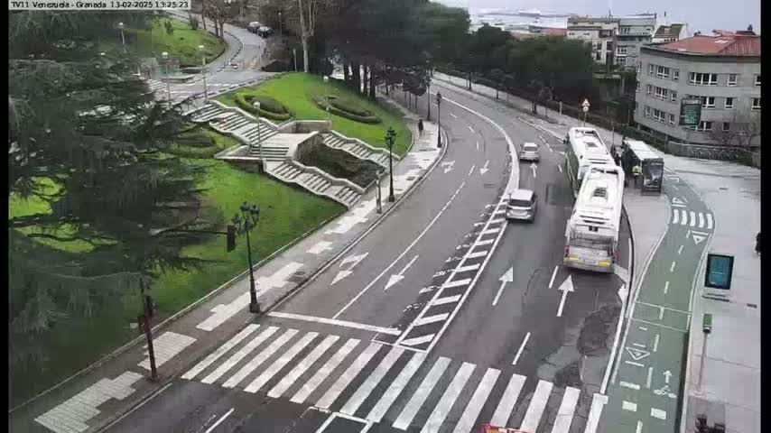 A curved road with a crosswalk and several cars and buses is seen on an overcast day, next to a grassy area with stairs and trees.