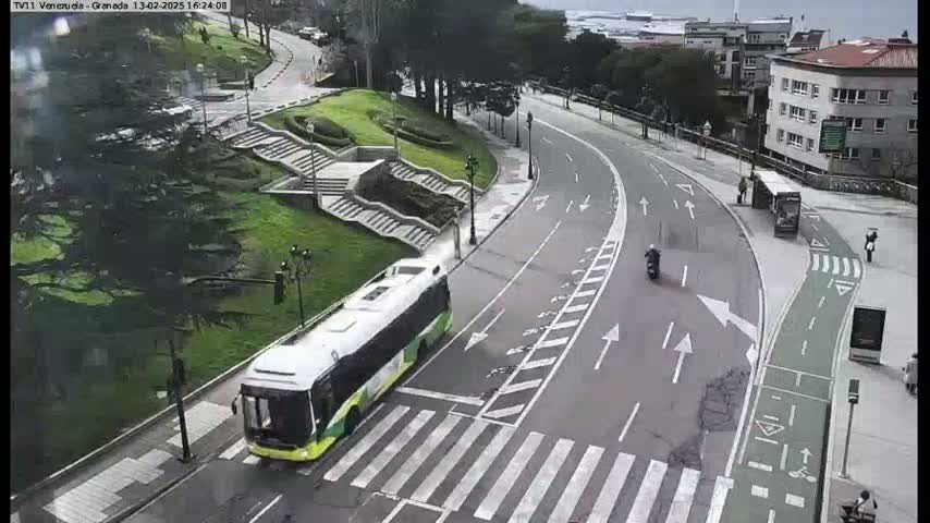 A bus and a motorcycle are traveling on a curved road next to a grassy area with stairs, under an overcast sky.