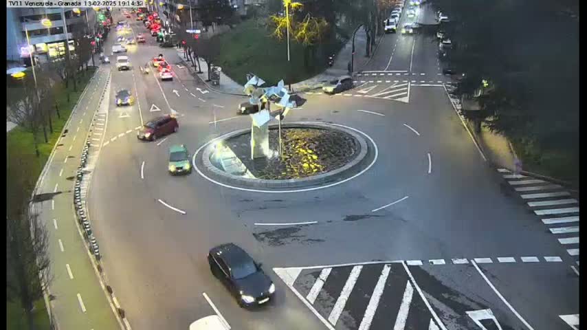 An aerial view shows a roundabout with several cars driving around it and several more cars on the roads leading to it at dusk.