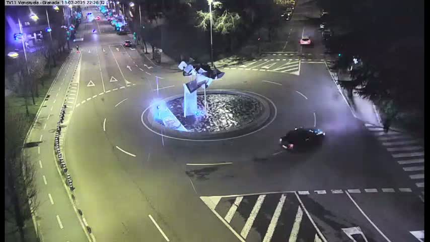 A nighttime aerial view shows a roundabout with a modern sculpture in the center, surrounded by a roadway with several cars and a pedestrian crossing.