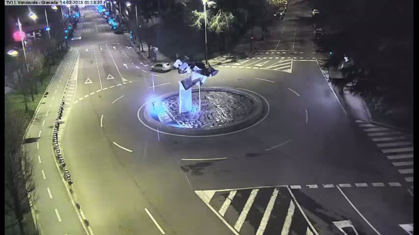 A nighttime aerial view shows a roundabout with a modern sculpture in the center, surrounded by roads with minimal traffic.