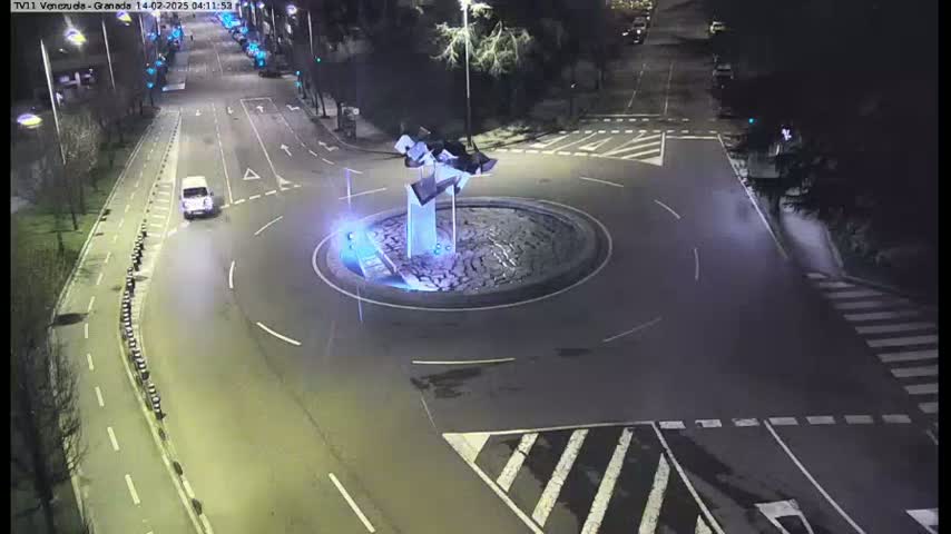 A nighttime aerial view shows a roundabout with a modern sculpture at its center, several cars navigating the roads around it, and pedestrian crosswalks visible on the surrounding streets.
