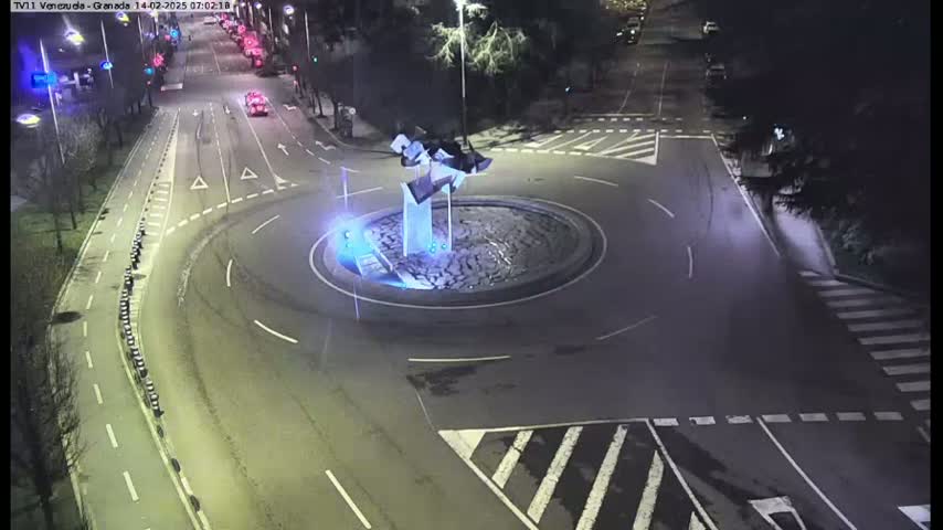 A nighttime aerial view of a roundabout with a modern sculpture in the center, surrounded by roads with cars and pedestrian crossings.