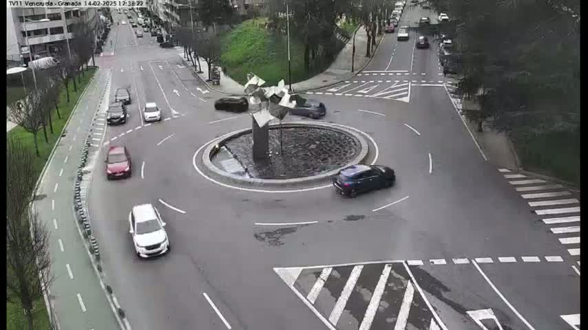 Several cars navigate a roundabout with a modern sculpture in the center, under an overcast sky.