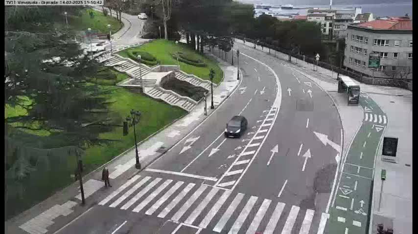 A curved road with a crosswalk and a bike lane winds through a park-like setting past a bus stop and several buildings under an overcast sky.