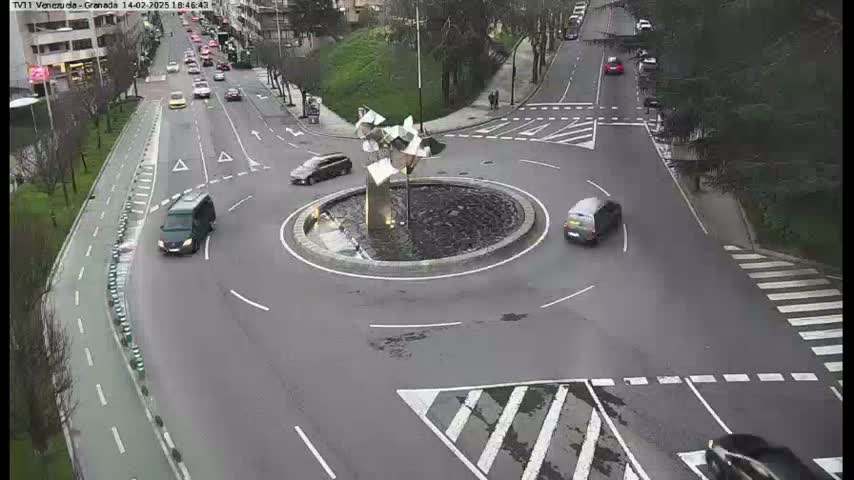 A roundabout with several cars navigating it, situated in a city with wet roads.