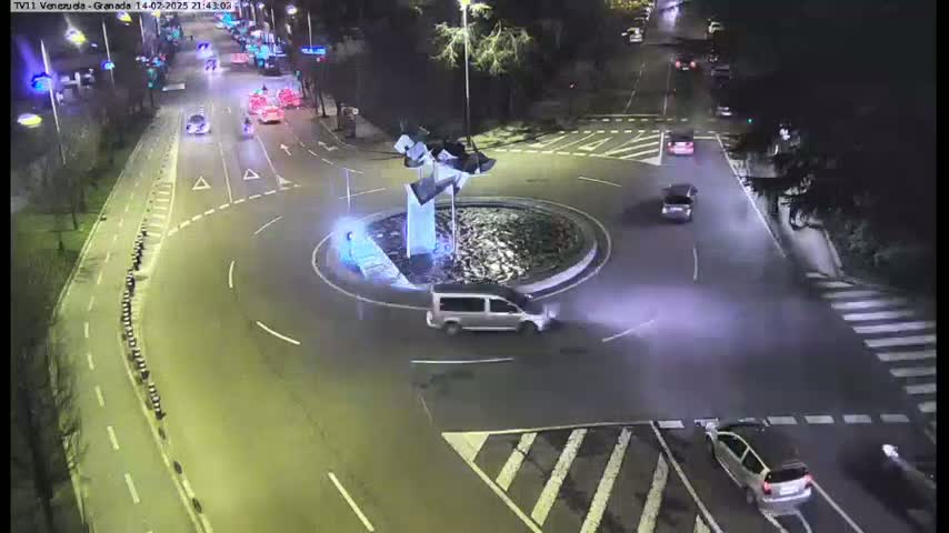 A nighttime aerial view shows several vehicles navigating a roundabout centered on a modern sculpture, with surrounding roads illuminated under artificial light.