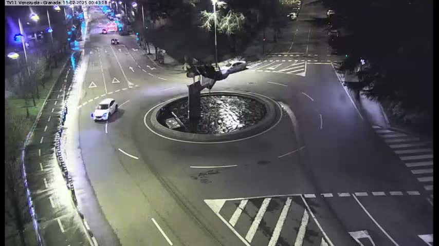 A nighttime aerial view shows a roundabout with a sculpture in the center, a few cars on nearby roads, and trees lining the streets.