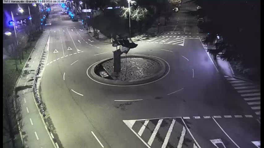 A nighttime aerial view of a roundabout with a dark sculpture in the center, surrounded by roads and crosswalks.