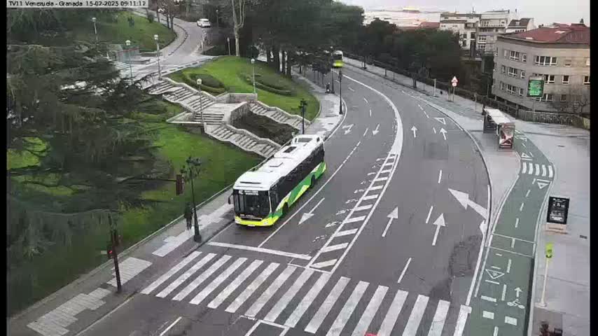 A green and white bus is stopped at a crosswalk on a curving road next to a grassy area with stairs, under an overcast sky.
