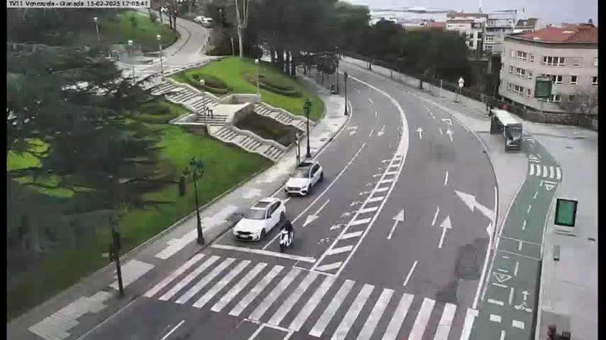 A curved road with a crosswalk and several cars, a motorcycle, and a bus, is situated next to a grassy area with stairs, all under an overcast sky.