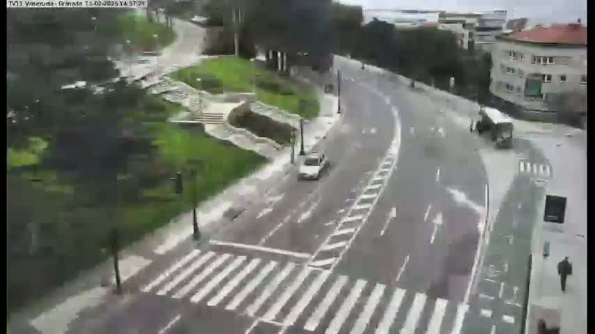 A light gray car drives on a wet, curved road that intersects a crosswalk near a pedestrian and a bus, all surrounded by lush green landscaping and buildings.