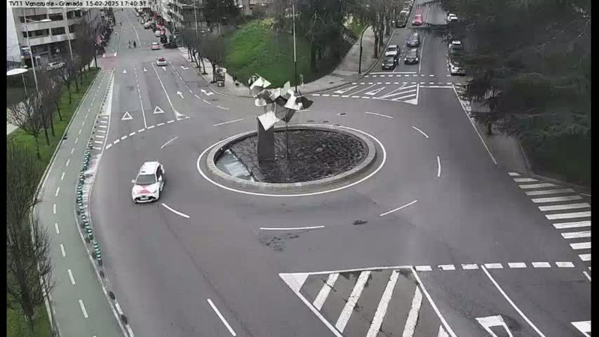A roundabout with a modern sculpture in the center is surrounded by roads with several cars, under a cloudy sky.