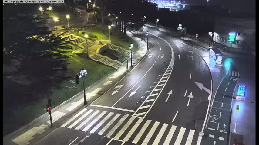 A nighttime view of a mostly empty roadway curving through a city park, illuminated by streetlights, with a pedestrian crossing and stairs visible.
