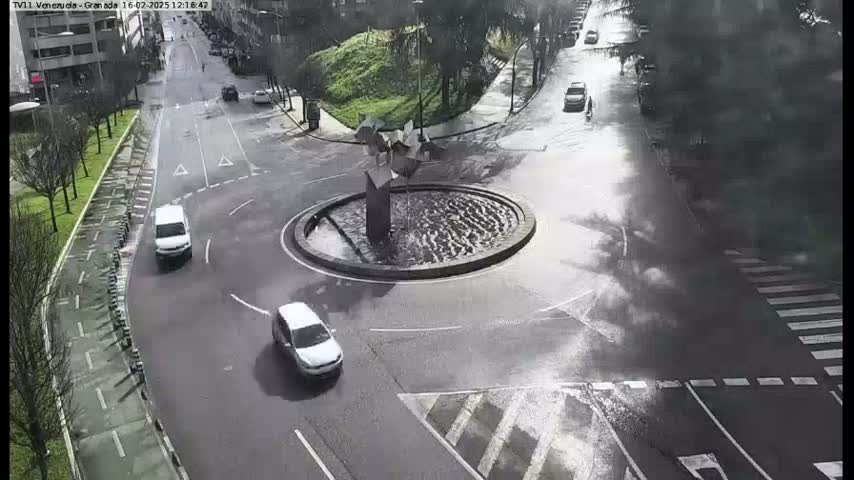 A circular traffic roundabout with a fountain in the center, featuring several cars and a pedestrian, on a wet, overcast day.