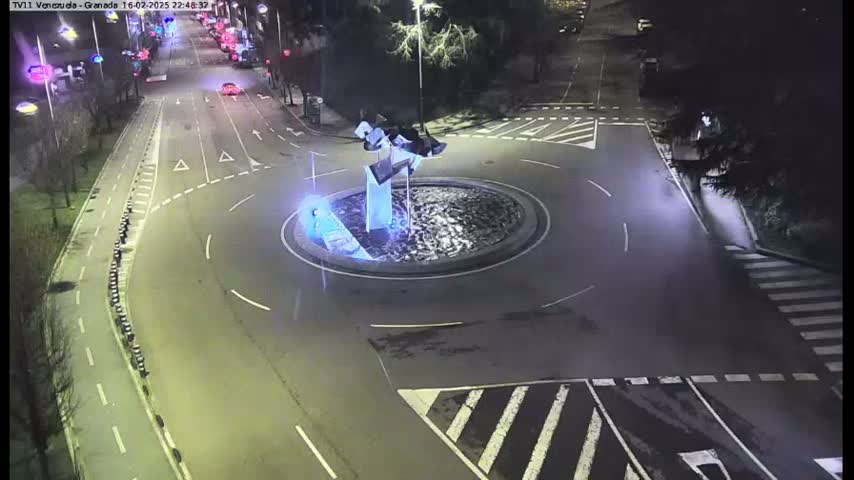 An overhead nighttime view shows a roundabout with an abstract sculpture in the center, surrounded by roads and pedestrian crossings.