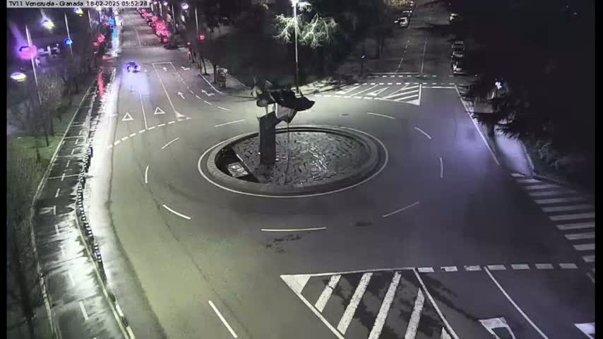A nighttime, wet road scene shows a roundabout with a sculpture in the center, surrounded by roads and crosswalks with minimal traffic.