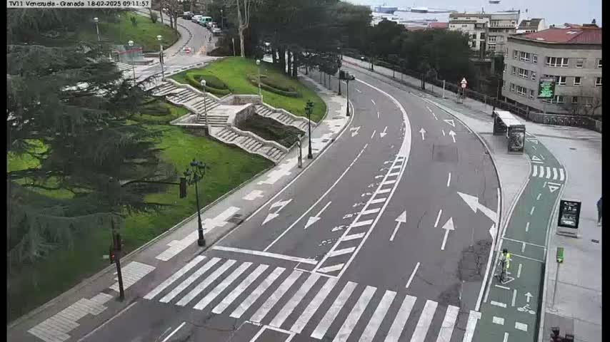 A mostly empty, curved roadway with pedestrian crosswalks and a bike lane winds through a green, landscaped area past stone steps and buildings under an overcast sky.