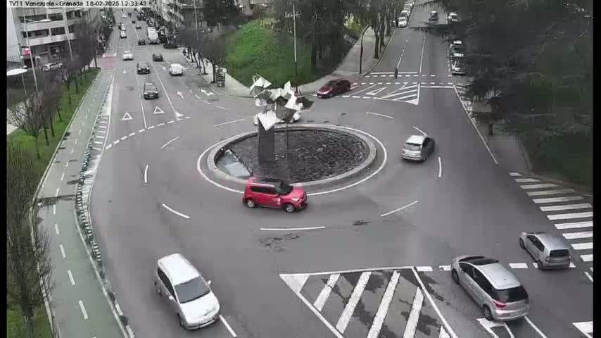 A roundabout with several cars driving around it and a modern art sculpture in the center, on a partly cloudy day.