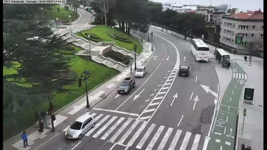 A curved road with several cars and buses, pedestrians crossing a crosswalk, and a grassy area with stairs in the background on a cloudy day.