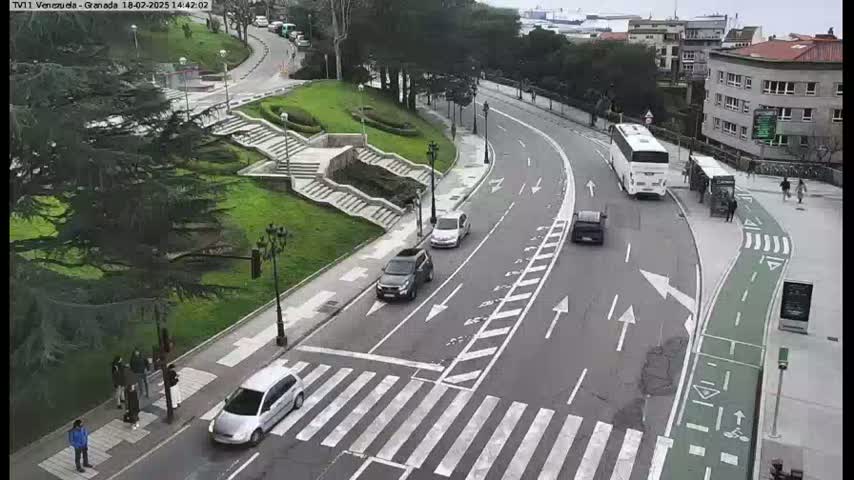 A curving road with several cars and a bus, pedestrians crossing at a crosswalk, and a grassy area with stairs in the background, under a cloudy sky.