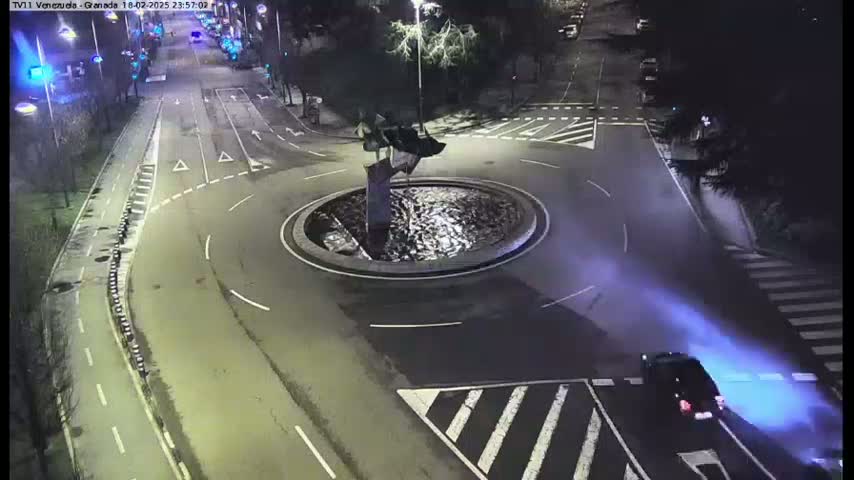 A dark, nighttime aerial view shows a roundabout with a central fountain, surrounded by roads with vehicles, and a car's headlights leaving a blue trail on the street.