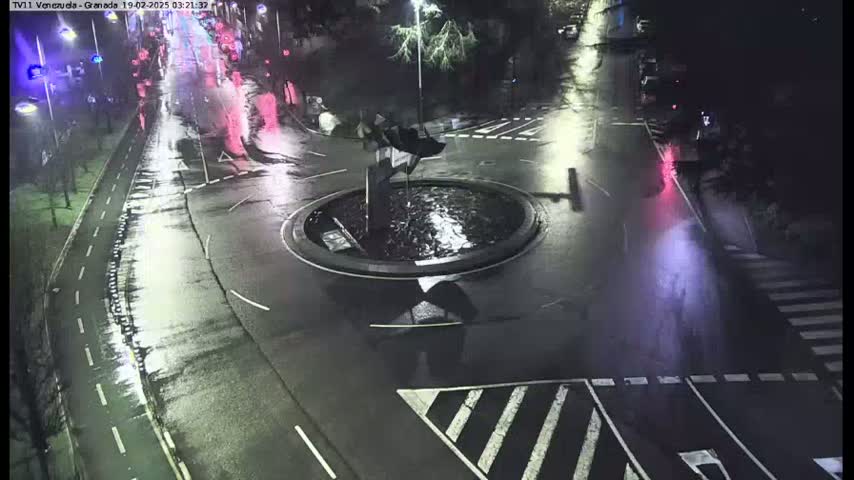 A wet, nighttime roundabout with sparse traffic is shown, centered around a small, dark fountain.