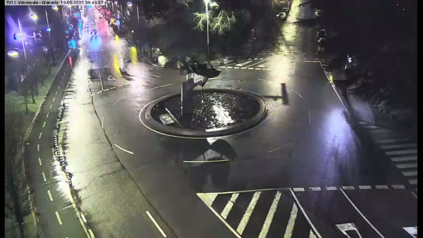 A wet, nighttime scene shows a roundabout with a fountain in the center, surrounded by roads and pedestrian crossings.
