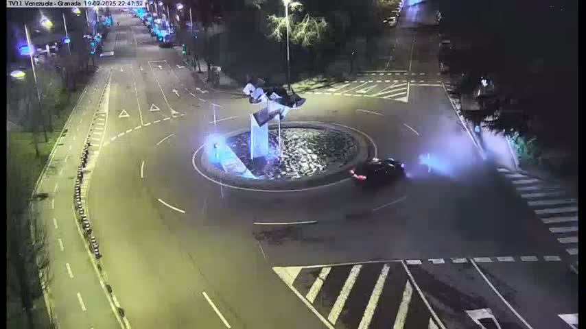 A nighttime aerial view of a roundabout with a single car in the center, surrounded by roads and streetlights.