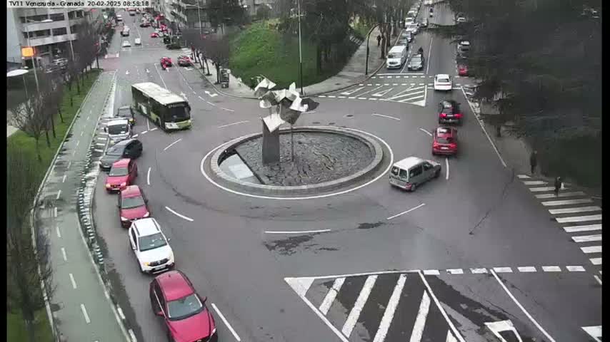 A roundabout with a modern sculpture in the center is surrounded by cars and a bus on a partly cloudy day.