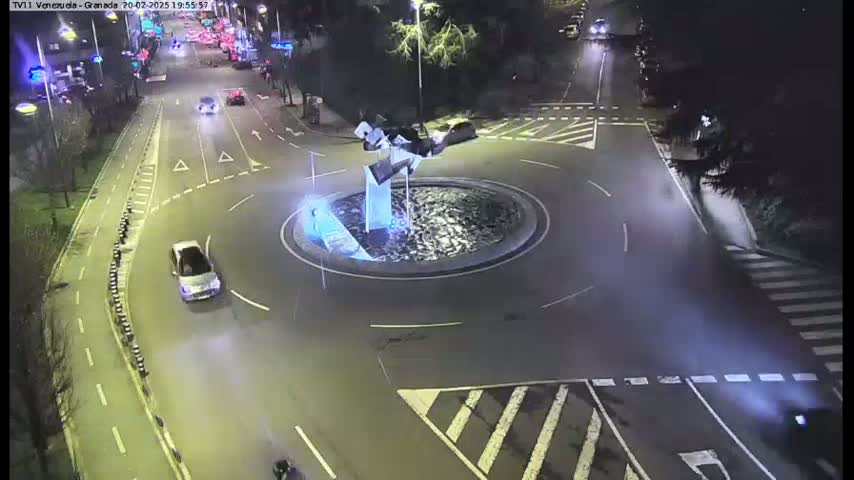 A nighttime aerial view shows a roundabout with a few cars, a modern sculpture in the center, and surrounding roads with pedestrian crossings.