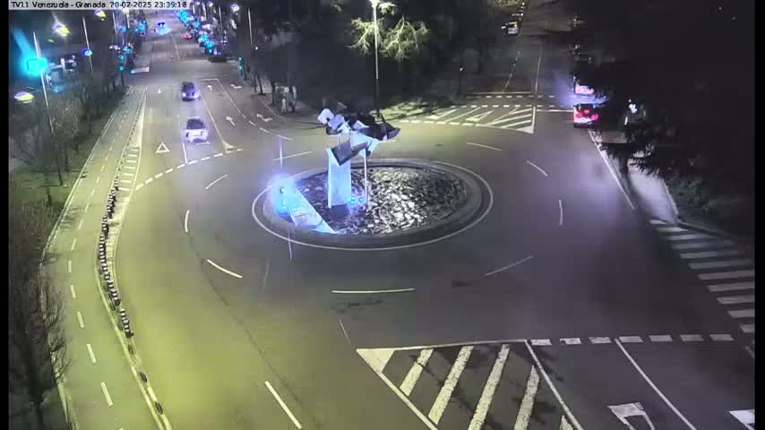 A nighttime aerial view shows a roundabout with a modern sculpture in the center, surrounded by roads with several cars.
