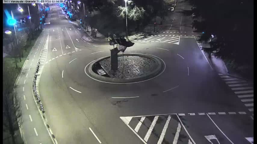 A nighttime aerial view shows a mostly empty roundabout with a dark sculpture in the center, surrounded by roads and crosswalks.