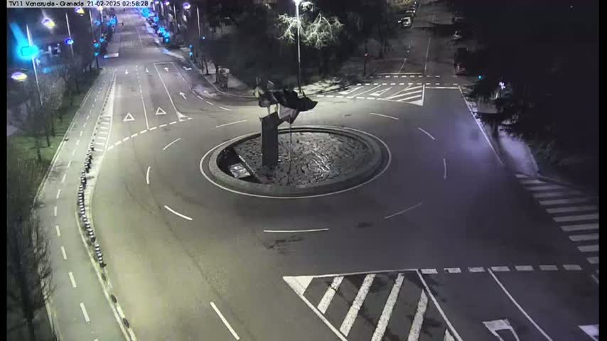 A nighttime aerial view shows a mostly empty roundabout with a dark sculpture in the center, surrounded by roads with faint lane markings.