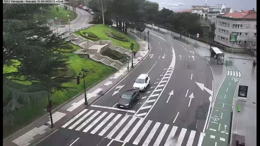 A wet road curves through a city area past a pedestrian crossing, with two cars and a bus visible near landscaped areas and buildings.