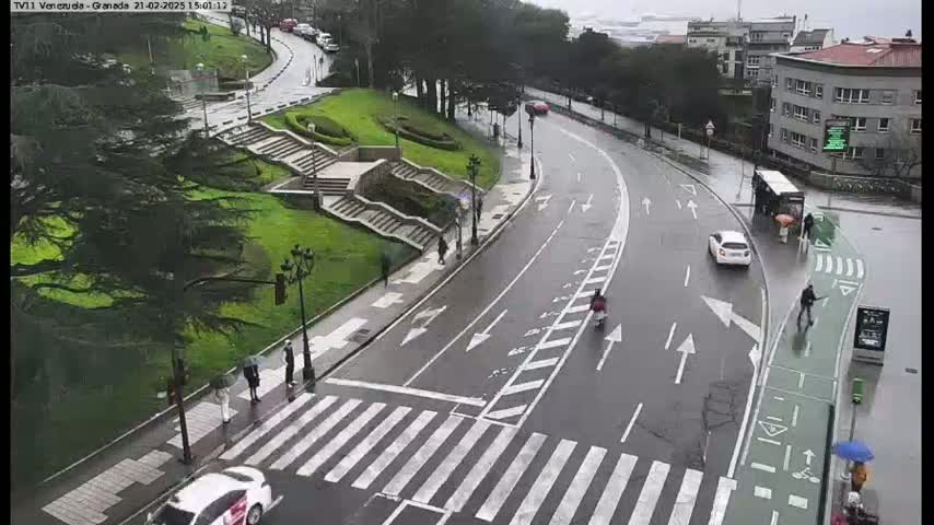 A wet, paved road curves through a city, past a pedestrian crossing, stairs leading to a grassy area with trees, a bus stop, and several people walking.