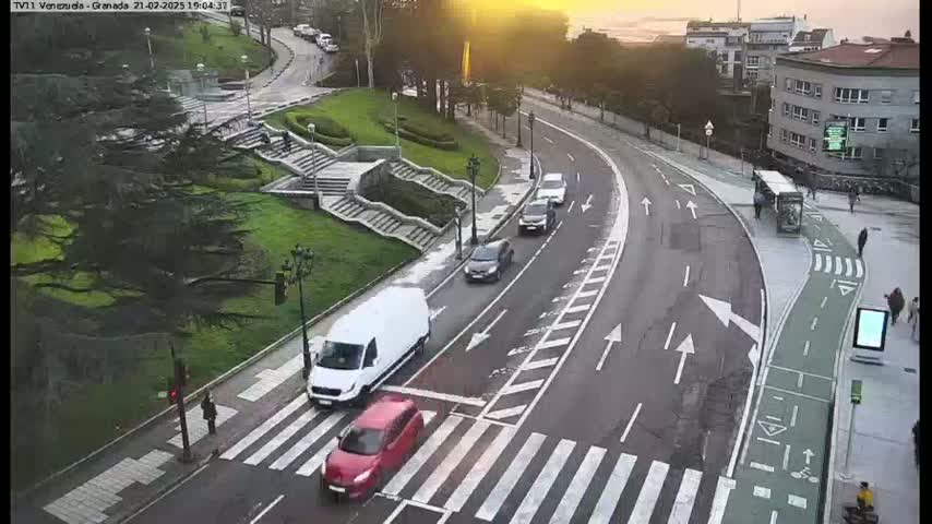 A curved road with several cars and a van, a pedestrian crossing, and a set of stairs leading down to a grassy area, all under a partly sunny sky.