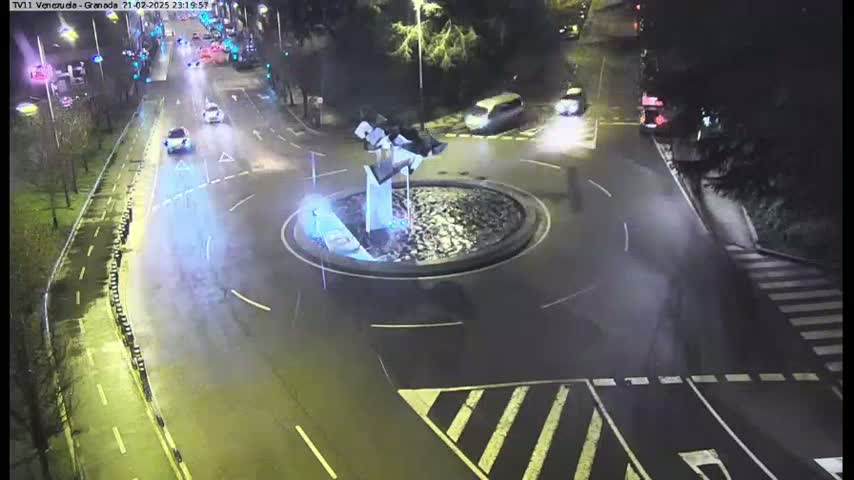 A wet, nighttime roundabout with a modern sculpture in the center and several cars driving on surrounding streets.