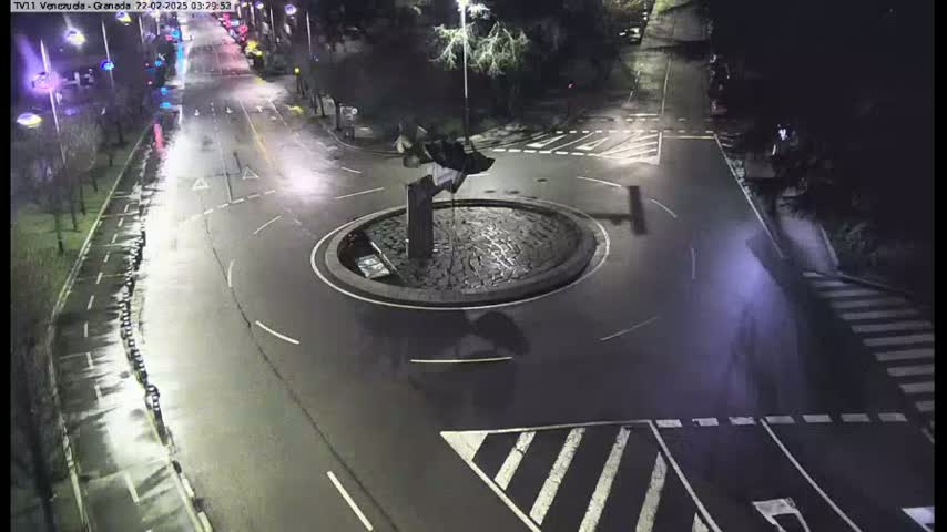 An overhead nighttime view shows a roundabout with a dark sculpture in the center, surrounded by wet, dark asphalt roads and pedestrian crossings.