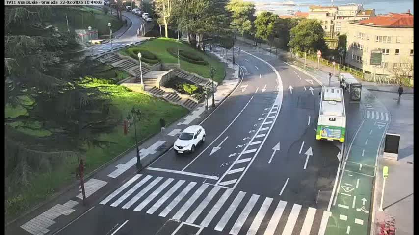 A wet road curves through a city area with a white car and a green bus, surrounded by green spaces and buildings, with pedestrians visible in the distance.
