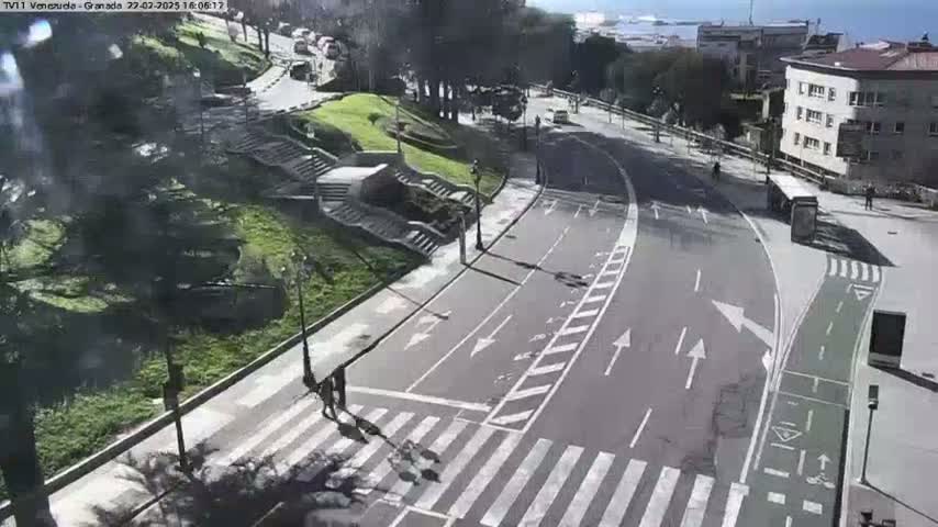 A sunny, overhead view of a paved road curving through a city park, with pedestrians crossing a crosswalk, and a bicycle lane alongside the road.