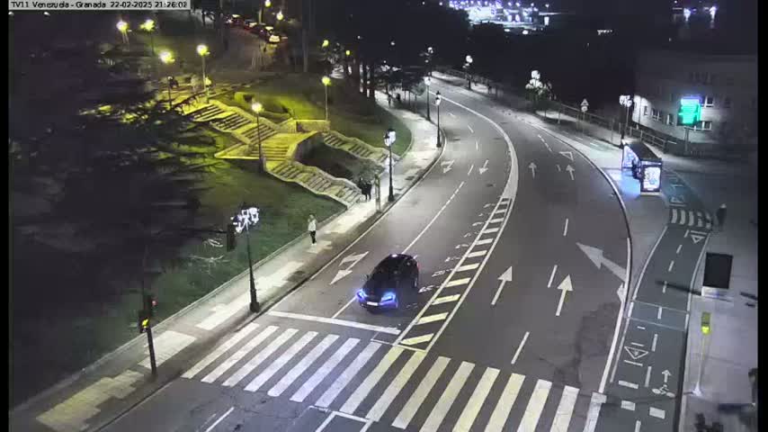 A dark-colored car drives on a well-lit road at night, passing a pedestrian crosswalk near stairs and trees.